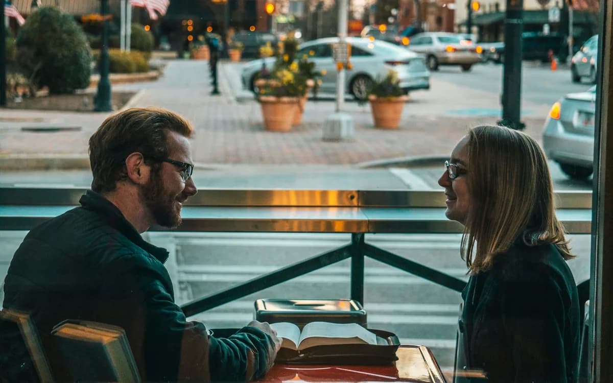 A couple at a cafe window table, facing each other mid-conversation, with no phones on the table